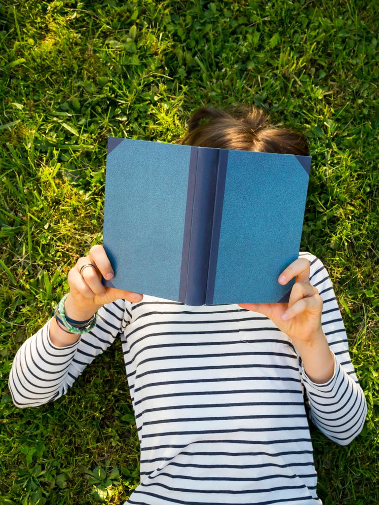 Ein Mädchen liegt auf einer Wiese und liest ein Buch. Ansicht von oben. Das Gesicht des Kindes ist unter dem Buch nicht zu erkennen.