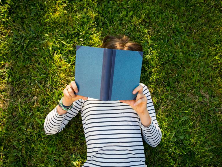 Ein Mädchen liegt auf einer Wiese und liest ein Buch. Ansicht von oben. Das Gesicht des Kindes ist unter dem Buch nicht zu erkennen. Ein Mädchen liegt auf einer Wiese und liest ein Buch. Ansicht von oben. Das Gesicht des Kindes ist unter dem Buch nicht zu erkennen.