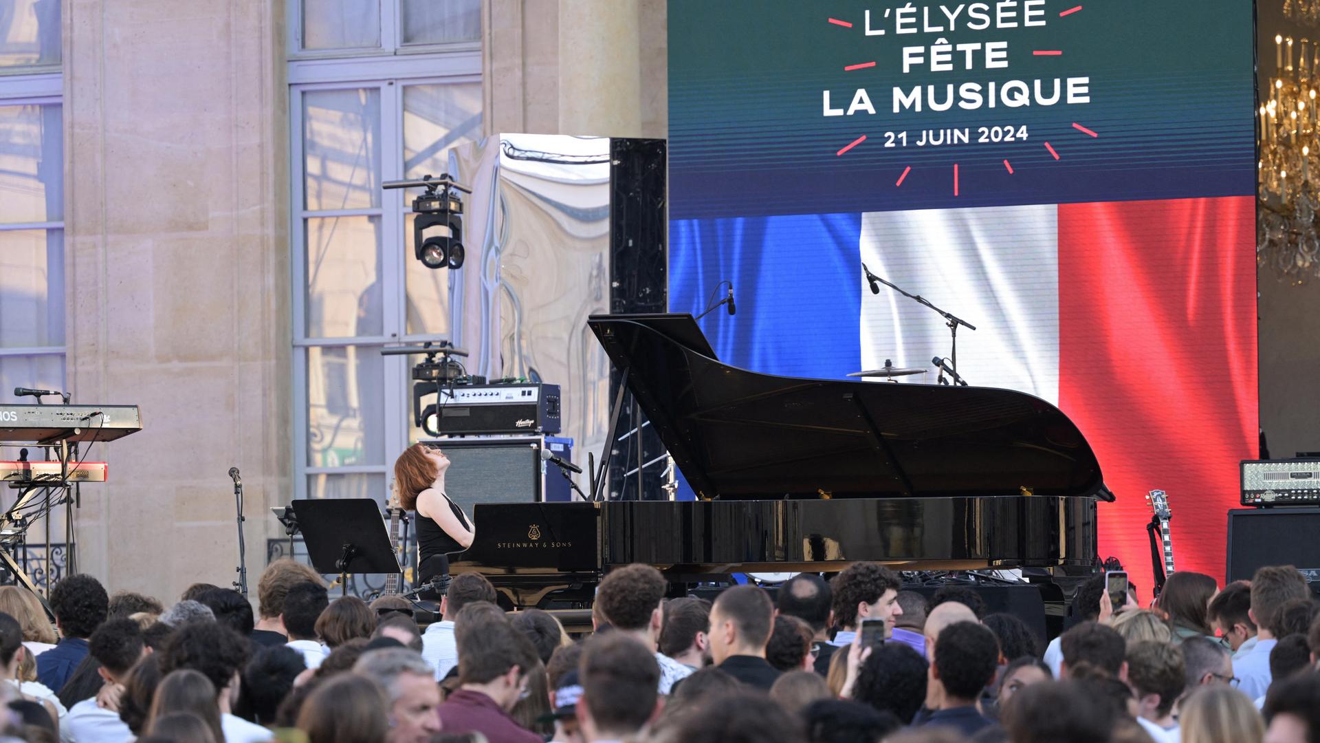 Eine Pianistin spielt vor der französischen Flagge, davor viele Menschen, dahinter eine Anzeige mit der Aufschrift "Fête de la musique" 