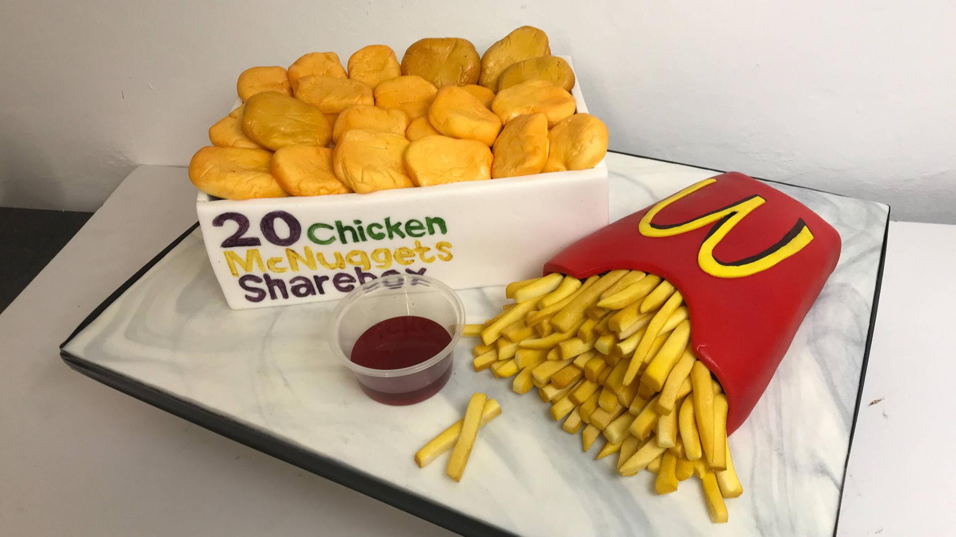 Eine Torte die einem Chicken-McNuggets-Menü einer Fastfood-Kette täuschend ähnlich sieht: Zu sehen sind, aus Teig gestaltet, eine Box mit Hähnchenstückchen und eine Portion Pommes Frite sowie ein Schälchen mit Sauce. Eine Torte die einem Chicken-McNuggets-Menü einer Fastfood-Kette täuschend ähnlich sieht: Zu sehen sind, aus Teig gestaltet, eine Box mit Hähnchenstückchen und eine Portion Pommes Frite sowie ein Schälchen mit Sauce.