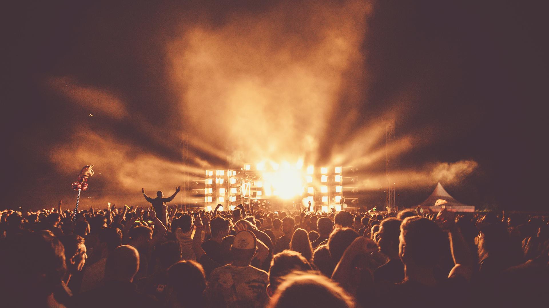 Menschenmengen tummeln sich vor einer beleuchteten Konzertbühne in gelblichem Licht.
