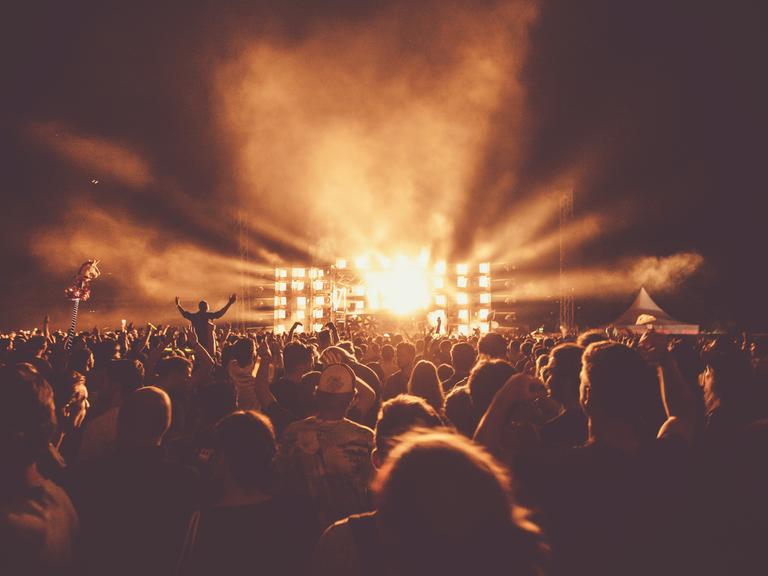 Menschenmengen tummeln sich vor einer beleuchteten Konzertbühne in gelblichem Licht.