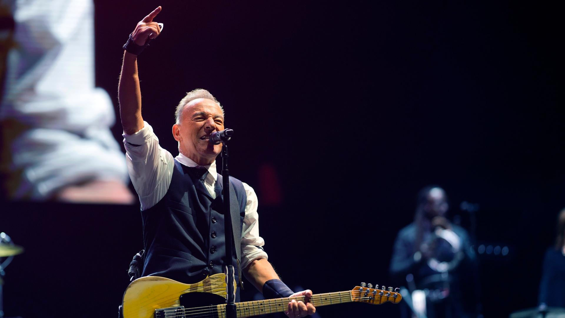 Der Rockmusiker Bruce Springsteen auf der Bühne beim Festival „Sea. Hear. Now“ in Asbury Park in New Jersey, USA. Er hat eine gelbe E-Gitarre umgehängt, singt ins Mikrofon und streckt zugleich den rechten Arm kraftvoll in die Luft. 