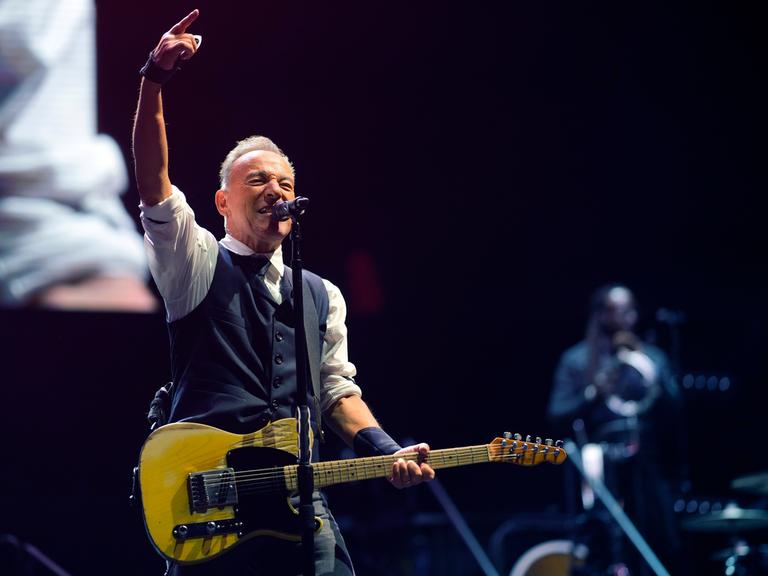 Der Rockmusiker Bruce Springsteen auf der Bühne beim Festival „Sea. Hear. Now“ in Asbury Park in New Jersey, USA. Er hat eine gelbe E-Gitarre umgehängt, singt ins Mikrofon und streckt zugleich den rechten Arm kraftvoll in die Luft. 