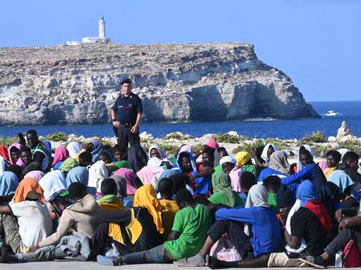 Lampedusa, Italien: Eine große Gruppe Migranten wartet in Cala Pisana darauf, von der Insel Lampedusa in Süditalien an andere Orte gebracht zu werden. Im Hintergrund ist ein Beamter zu sehen und ein Felsen mit einem Leuchtturm.