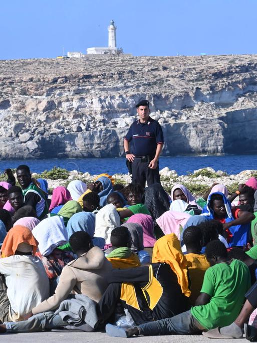 Lampedusa, Italien: Eine große Gruppe Migranten wartet in Cala Pisana darauf, von der Insel Lampedusa in Süditalien an andere Orte gebracht zu werden. Im Hintergrund ist ein Beamter zu sehen und ein Felsen mit einem Leuchtturm.