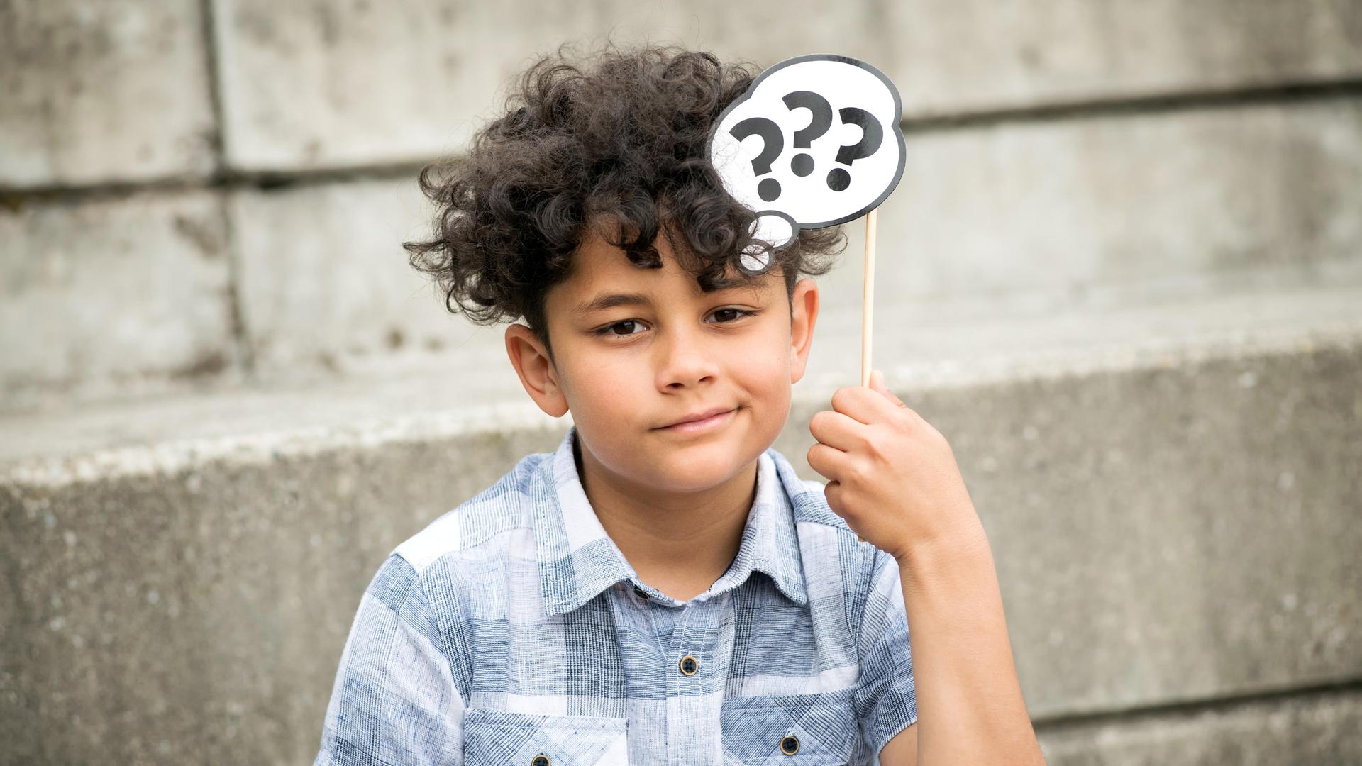 Ein Junge denkt nach und hält über seinen Kopf eine Gedankenblase aus Papier mit Fragezeichen. 