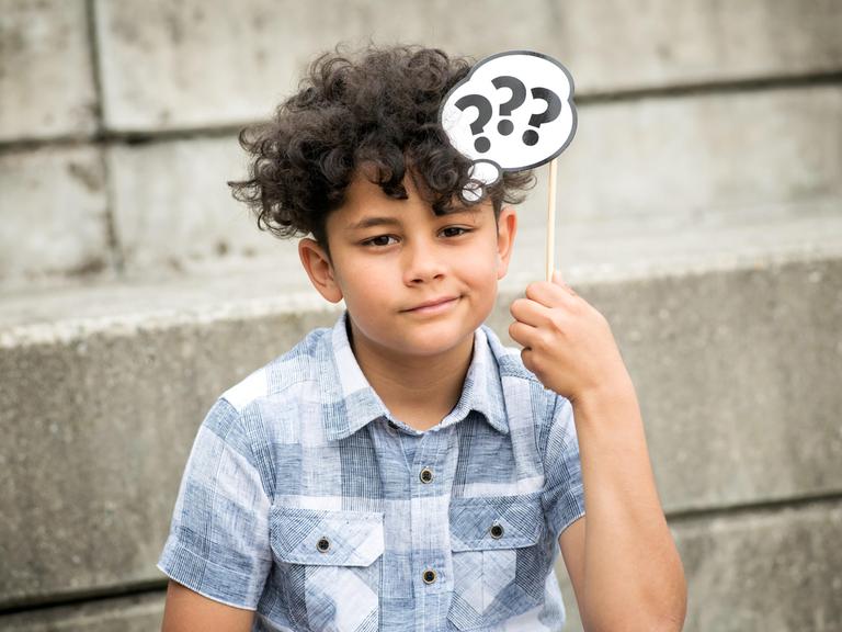 Ein Junge denkt nach und hält über seinen Kopf eine Gedankenblase aus Papier mit Fragezeichen. 