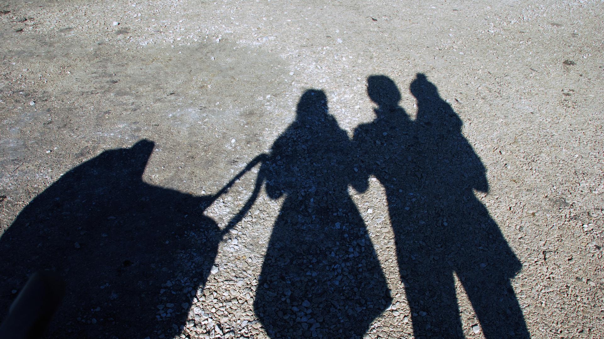 Die Schatten von Personen fallen auf eine Straße; zu erkennen sind zwei Personen und ein Kind, ebenso ein Kinderwagen.