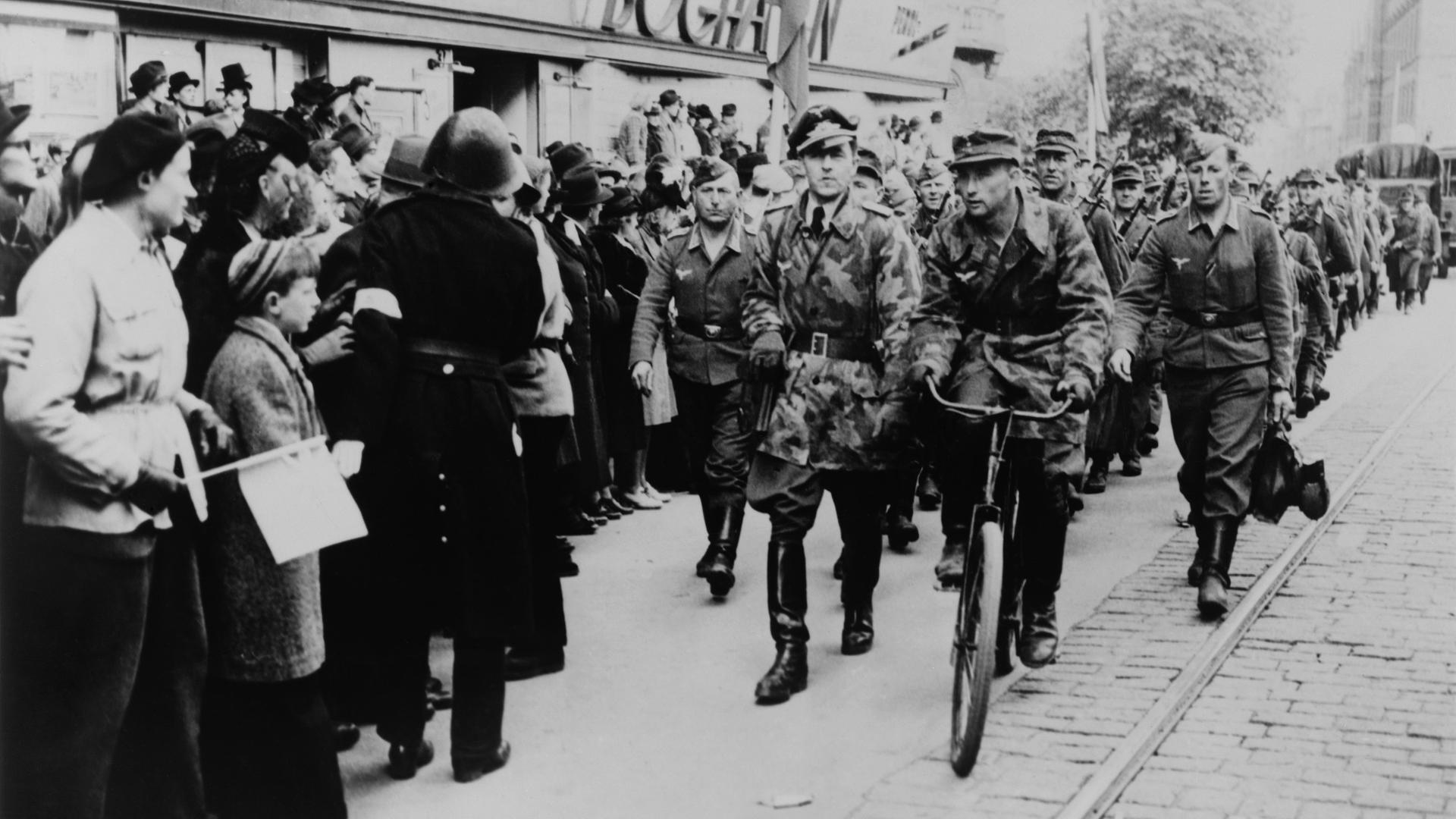 Nazi-Soldaten marschieren 1945 eine Geschäftsstraße im deutsch besetzten Dänemark entlang. Am Straßenrand steht die Bevölkerung.