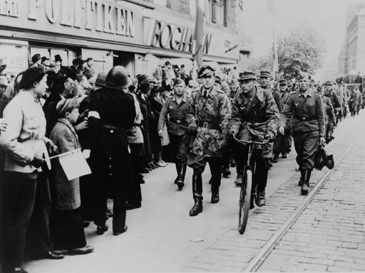 Nazi-Soldaten marschieren 1945 eine Geschäftsstraße im deutsch besetzten Dänemark entlang. Am Straßenrand steht die Bevölkerung.