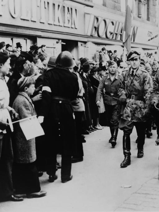 Nazi-Soldaten marschieren 1945 eine Geschäftsstraße im deutsch besetzten Dänemark entlang. Am Straßenrand steht die Bevölkerung.