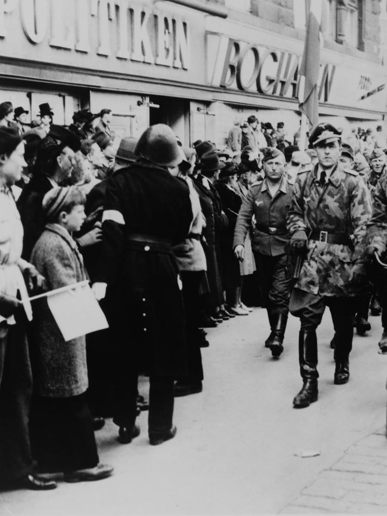 Nazi-Soldaten marschieren 1945 eine Geschäftsstraße im deutsch besetzten Dänemark entlang. Am Straßenrand steht die Bevölkerung.