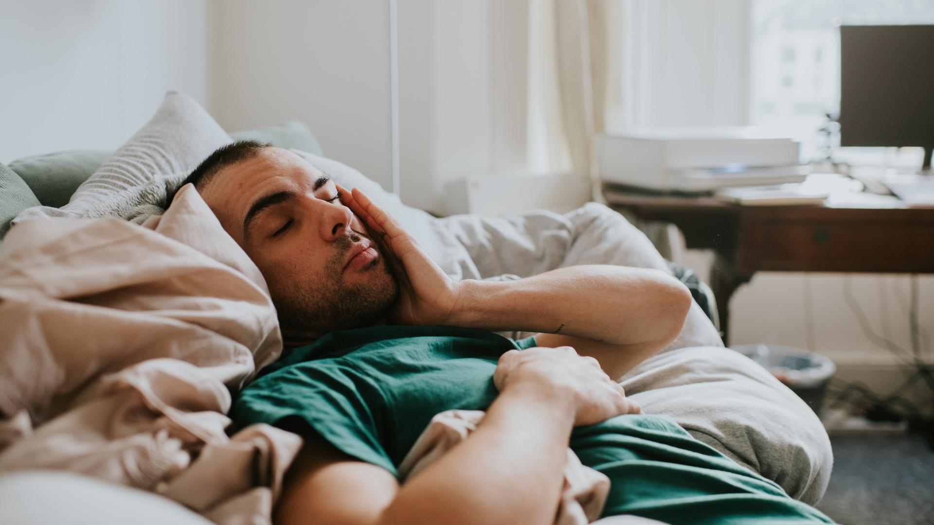 Ein Mann liegt zugedeckt in einem Bett und reibt sich mit der Hand über das Gesicht. Er sieht erschöpft aus.