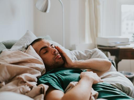 Ein Mann liegt zugedeckt in einem Bett und reibt sich mit der Hand über das Gesicht. Er sieht erschöpft aus.