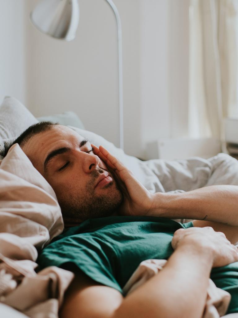 Ein Mann liegt zugedeckt in einem Bett und reibt sich mit der Hand über das Gesicht. Er sieht erschöpft aus.