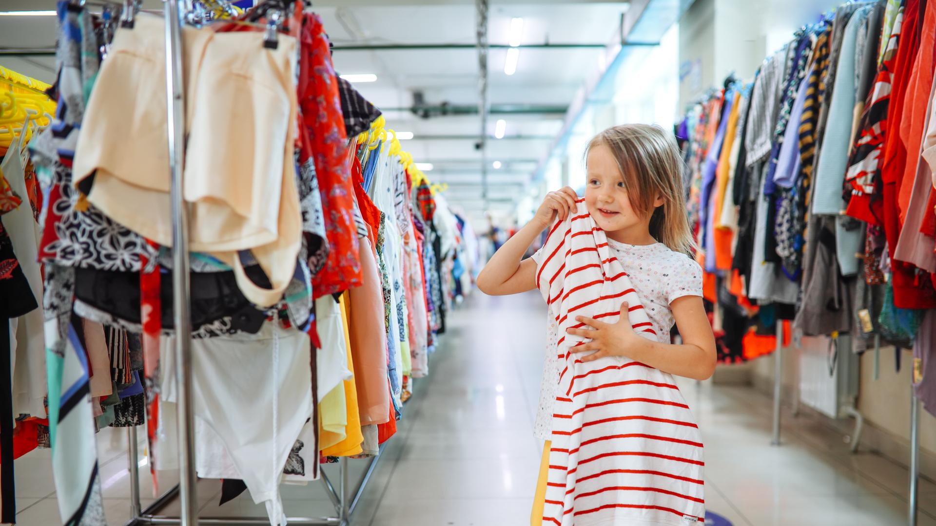 Ein Mädchen hält ein Kleid in einem Second-Hand Store, sie steht zwischen langen Reihen mit Kleidung. 