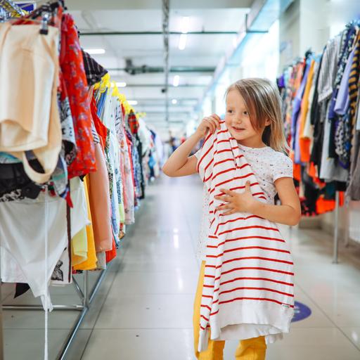 Ein Mädchen hält ein Kleid in einem Second-Hand Store, sie steht zwischen langen Reihen mit Kleidung. 