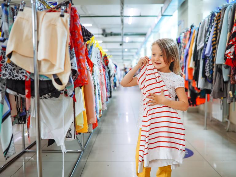 Ein Mädchen hält ein Kleid in einem Second-Hand Store, sie steht zwischen langen Reihen mit Kleidung. 