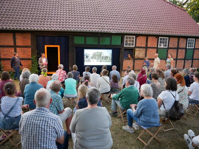 Eine Gruppe von Menschen sitzt auf Klappstühlen vor einem Fachwerkhaus und betrachtet die Projektion auf einem Bildschirm.
