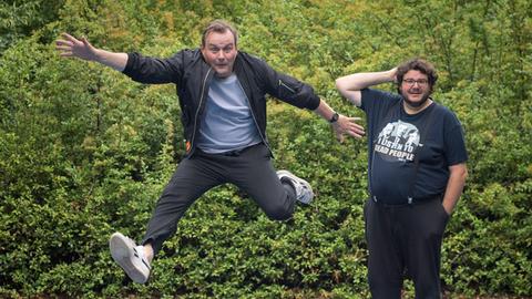 Devid Striesow und Axel Ranisch stehen auf einem Spielplatz mit Bodentrampolin. Devid Striesow springt in die Luft.