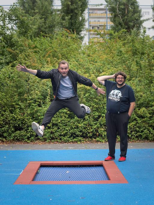 Devid Striesow und Axel Ranisch stehen auf einem Spielplatz mit Bodentrampolin. Devid Striesow springt in die Luft.
