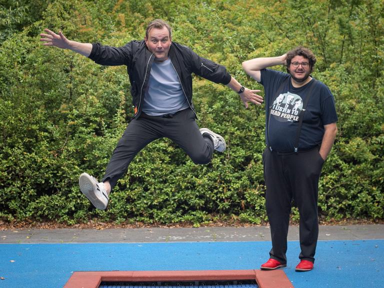 Devid Striesow und Axel Ranisch stehen auf einem Spielplatz mit Bodentrampolin. Devid Striesow springt in die Luft.
