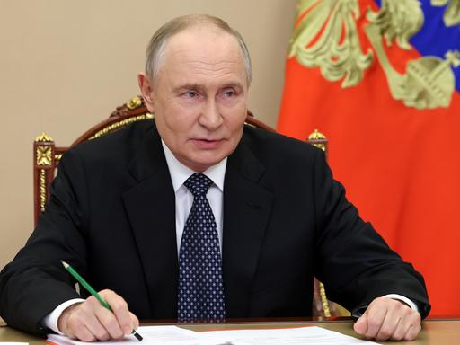 Russlands Präsident Wladimir Putin sitzt bei einem Treffen an einem Tisch, beide Unterarme aufgestützt, in der linken Hand einen Bleistift mit der Spitze nach oben.