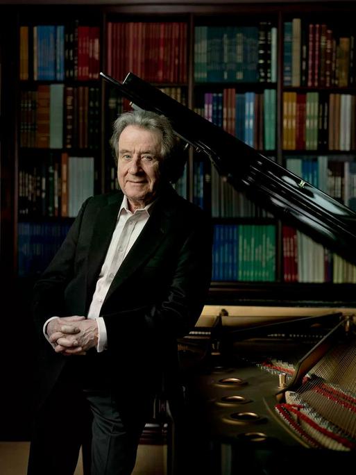 Rudolf Buchbinder steht an seinem heimischen Flügel, hinter dem Regale voller Noten zu erkennen sind.