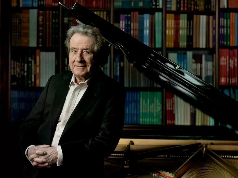 Rudolf Buchbinder steht an seinem heimischen Flügel, hinter dem Regale voller Noten zu erkennen sind.