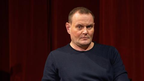 Der Schauspieler Devid Striesow sitzt mit ernstem Blick auf einer Lesebühne