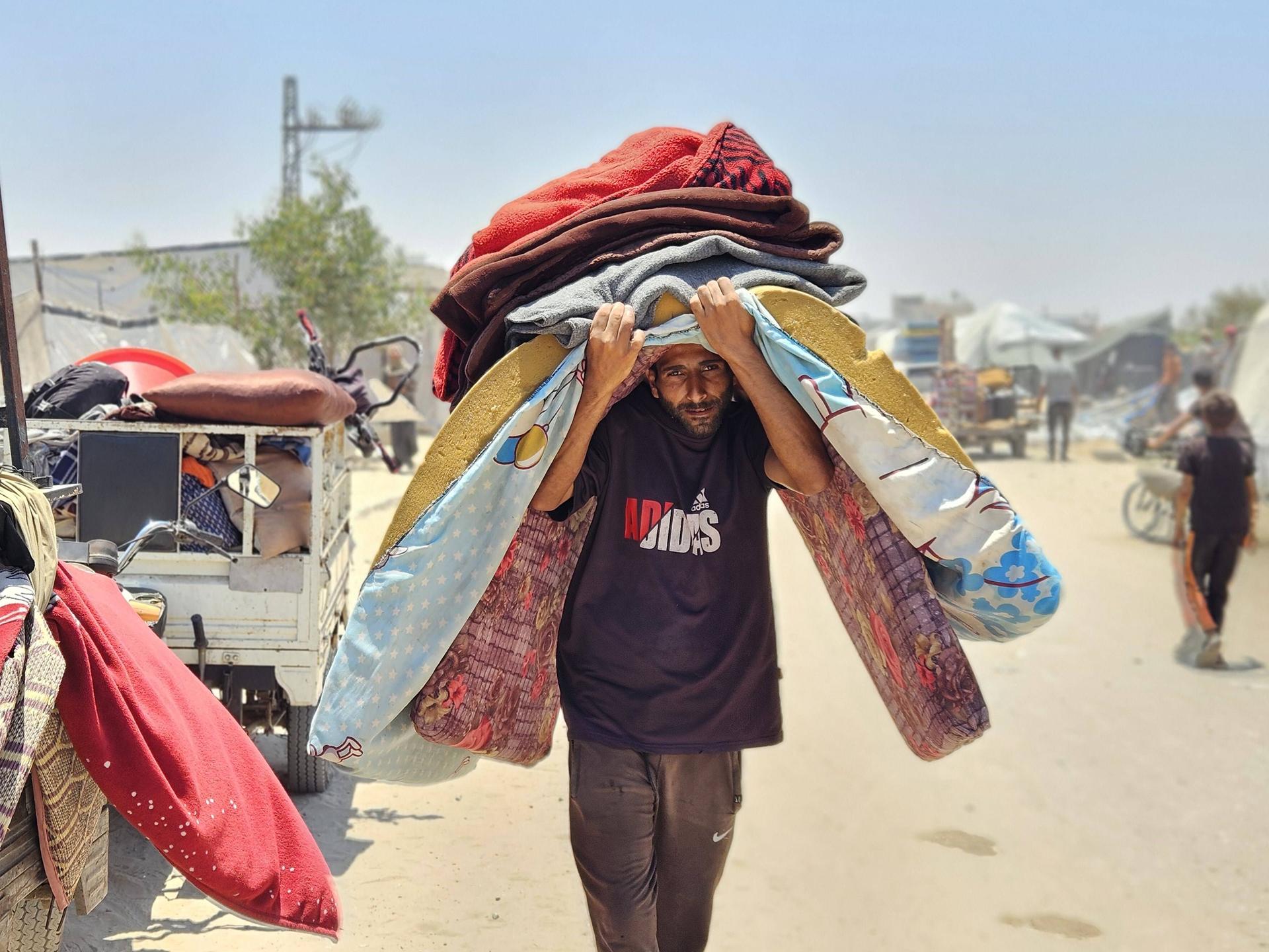 Ein Mann im Gazastreifen geht eine staubige Straße entlang. Er geht Richtung Kamera und trägt mehrere Gegenstände auf dem Kopf, darunter eine Matratze. Er geht gebeugt und ist sichtbar belastet. 