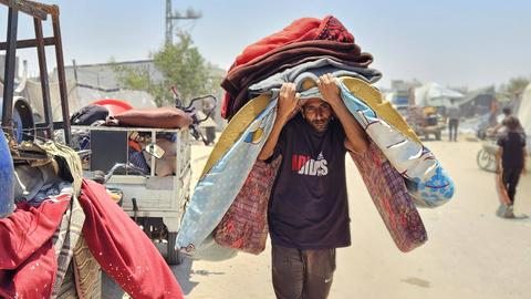 Ein Mann im Gazastreifen geht eine staubige Straße entlang. Er geht Richtung Kamera und trägt mehrere Gegenstände auf dem Kopf, darunter eine Matratze. Er geht gebeugt und ist sichtbar belastet. 