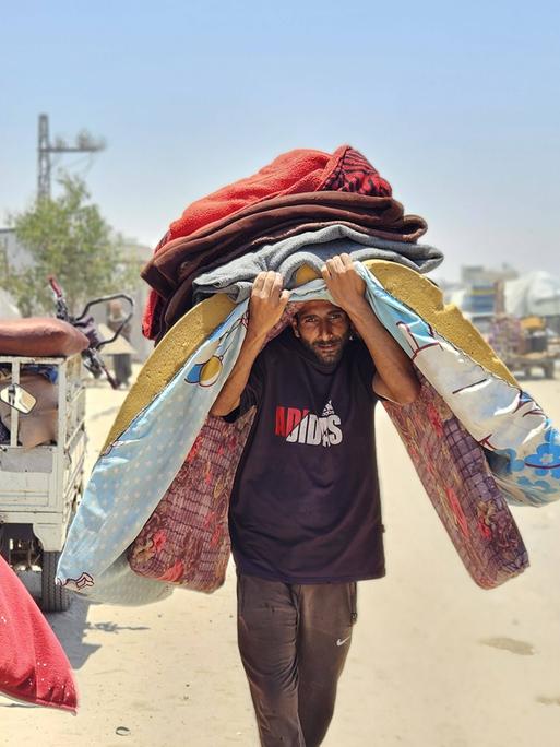 Ein Mann im Gazastreifen geht eine staubige Straße entlang. Er geht Richtung Kamera und trägt mehrere Gegenstände auf dem Kopf, darunter eine Matratze. Er geht gebeugt und ist sichtbar belastet. 