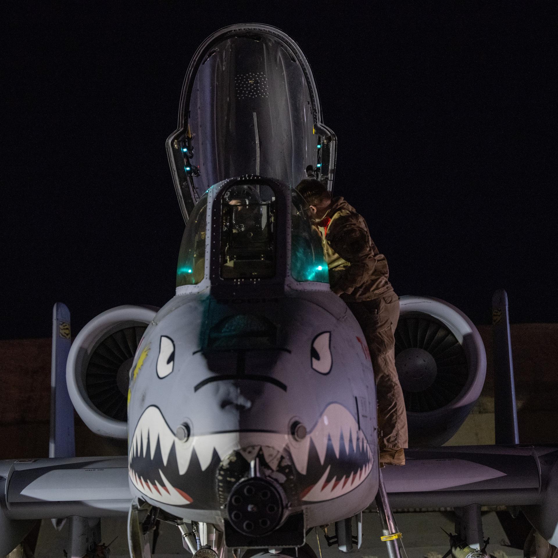 Ein US-Soldat bereitet einen Kampfjet auf den Flug vor. Es ist Nacht. Der Vorderteil des Flugzeugs ist mit einem Hai-Gesicht bemalt.