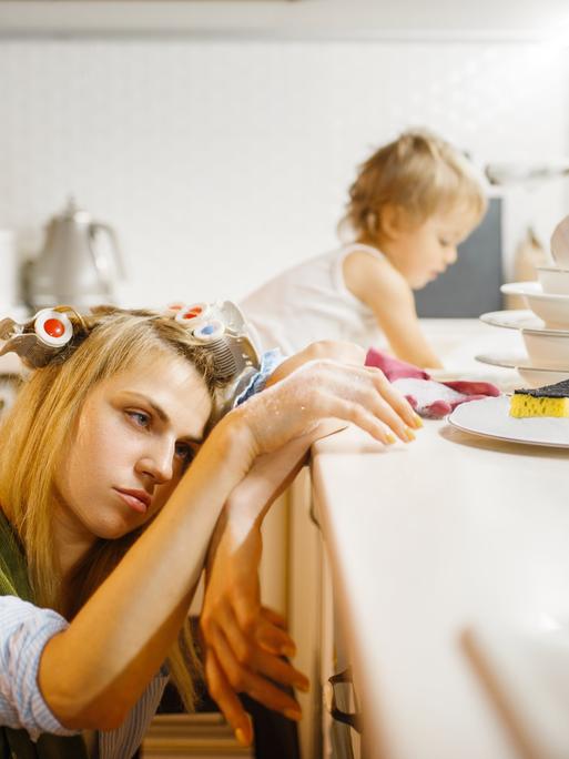 Eine Frau erschöpft - ihr Kopf liegt auf der Küchenarbeitsplatte. Ihr Kind turnt in der Spüle. 