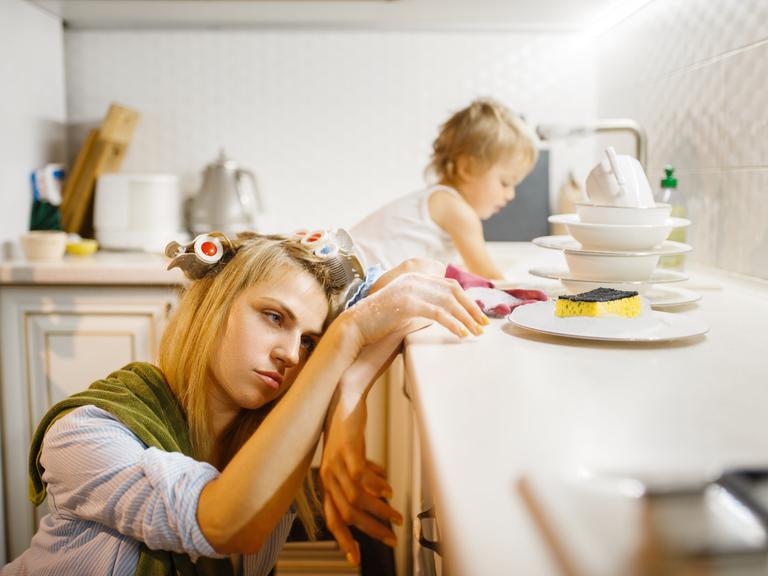Eine Frau erschöpft - ihr Kopf liegt auf der Küchenarbeitsplatte. Ihr Kind turnt in der Spüle. 