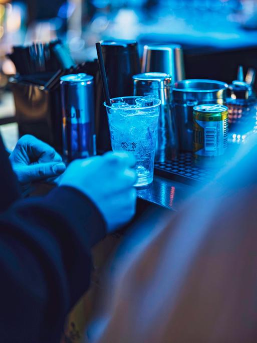 In einem Nachtclub stehen im blauen Licht Gläser und Dosen auf einem Tresen, davor stehen mehrere Menschen