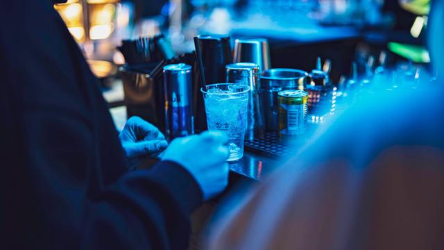 In einem Nachtclub stehen im blauen Licht Gläser und Dosen auf einem Tresen, davor stehen mehrere Menschen