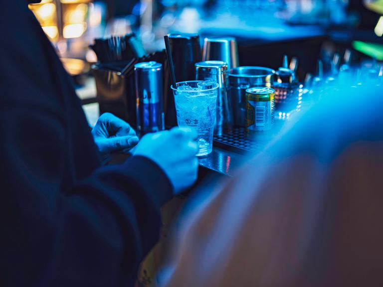 In einem Nachtclub stehen im blauen Licht Gläser und Dosen auf einem Tresen, davor stehen mehrere Menschen