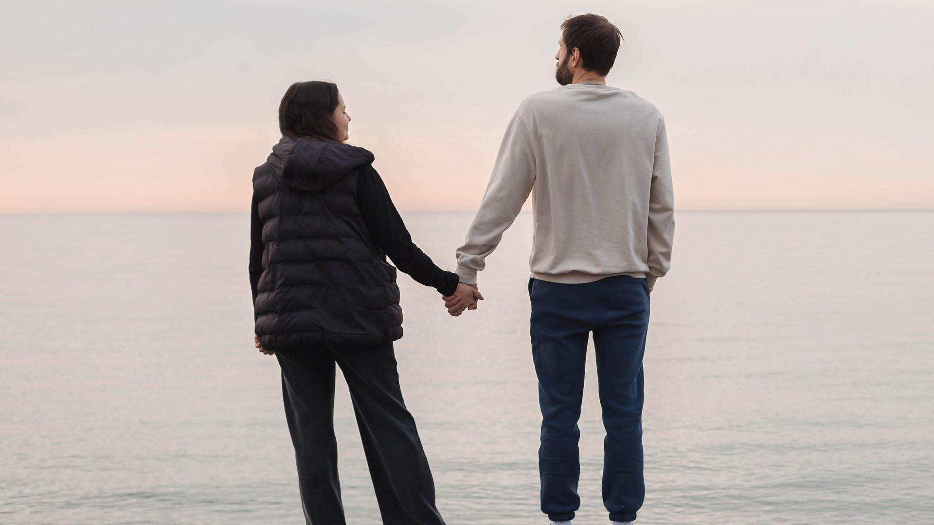 Ein Paar steht an einem Strand und hält sich an den Händen beim Blick aufs Meer (Symbolfoto).