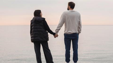 Ein Paar steht an einem Strand und hält sich an den Händen beim Blick aufs Meer (Symbolfoto). Ein Paar steht an einem Strand und hält sich an den Händen beim Blick aufs Meer (Symbolfoto).