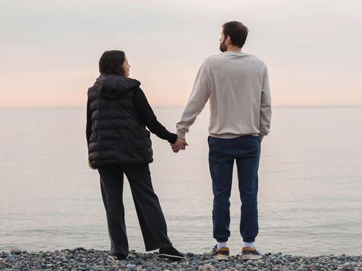 Ein Paar steht an einem Strand und hält sich an den Händen beim Blick aufs Meer (Symbolfoto). Ein Paar steht an einem Strand und hält sich an den Händen beim Blick aufs Meer (Symbolfoto).