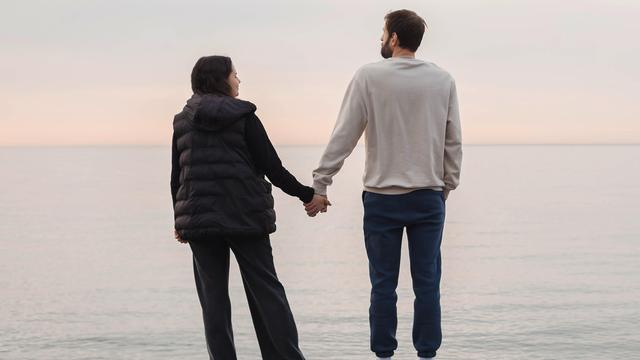 Ein Paar steht an einem Strand und hält sich an den Händen beim Blick aufs Meer (Symbolfoto).