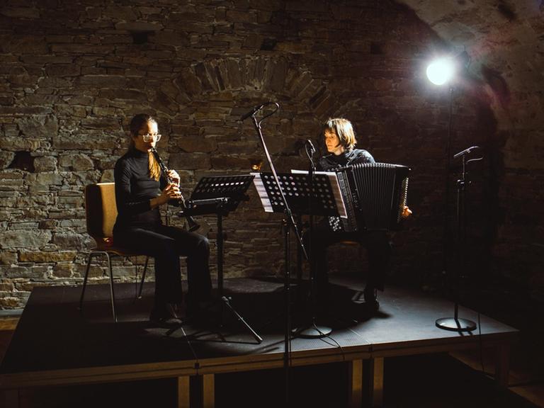 Auf dem Podium eines Konzertkellers sitzen zwei Instrumentalistinnen mit Klarinette und Akkordeon im Scheinwerferlicht