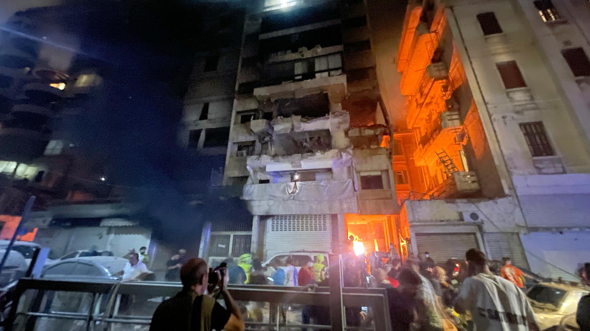 Ein Haus liegt in Trümmer. Ein feuer lodert. Menschen stehen um die Ruine herum.