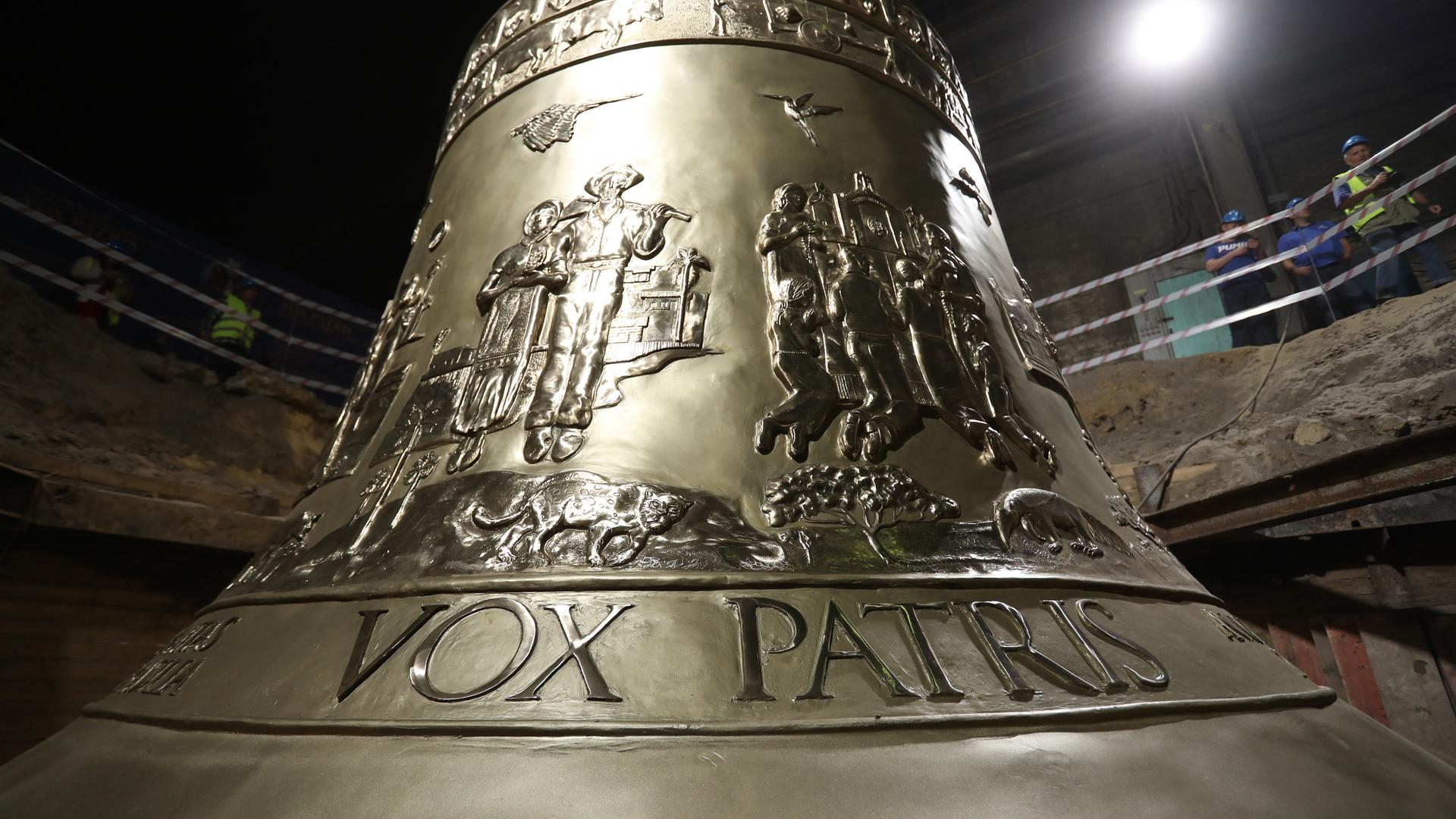 Zu sehen ist eine Glocke aus dunklem Metall mit goldenem Schimmer. Auf ihr sind Menschen und ein Tier zu sehen, unten steht in großen Buchstaben "Vox Patris". Zu sehen ist eine Glocke aus dunklem Metall mit goldenem Schimmer. Auf ihr sind Menschen und ein Tier zu sehen, unten steht in großen Buchstaben "Vox Patris".