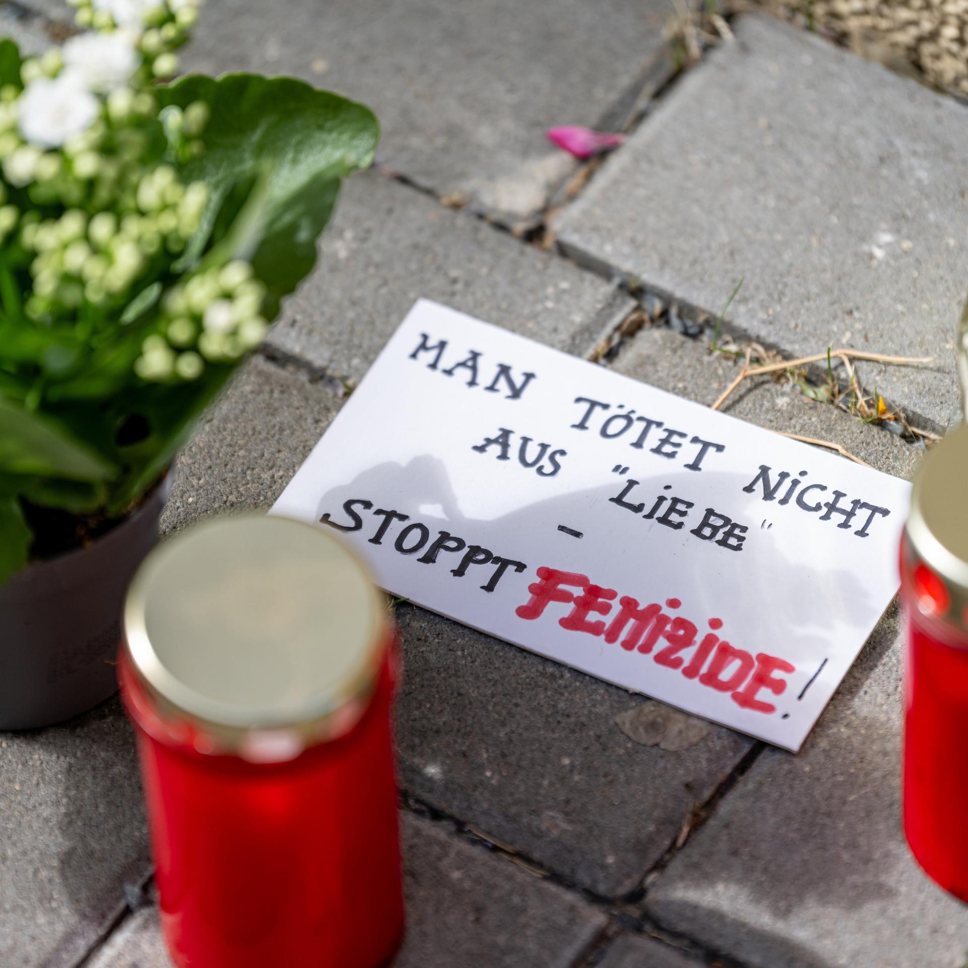 Blumen und Kerzen liegen nach einem tödlichen Messerangriff vor einem Hauseingang in Berlin-Friedrichsfelde neben einem Zettel mit der Aufschrift „Man tötet nicht aus Liebe - Stoppt Femizide“.