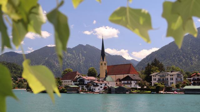 Blick auf den Tegernsee und die Kirche St.Laurentius in Rottach-Egern an einem Sommertag mit Blättern umrahmt.