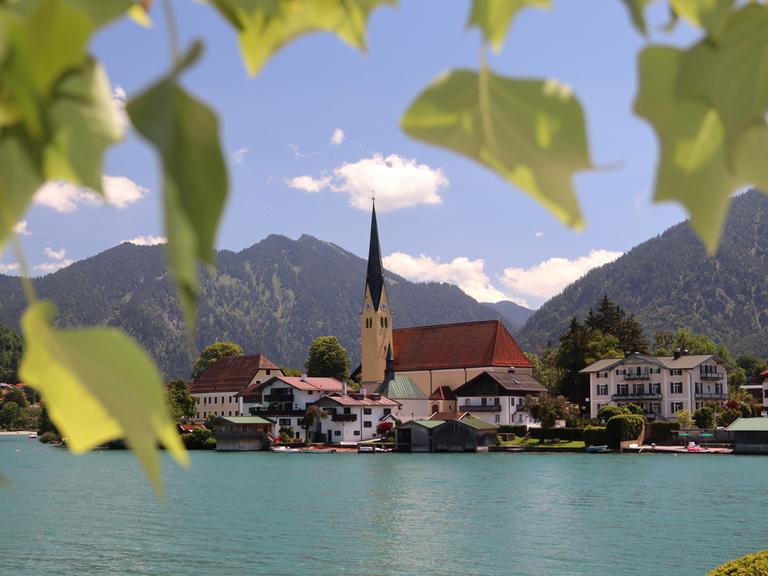 Blick auf den Tegernsee und die Kirche St.Laurentius in Rottach-Egern an einem Sommertag mit Blättern umrahmt.